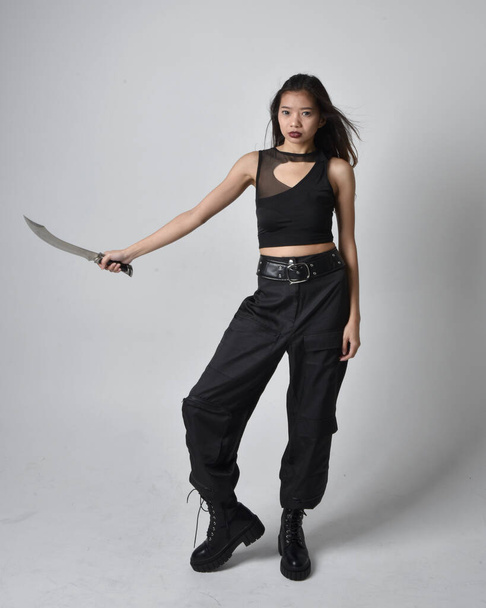 Full length portrait of pretty young asian girl wearing black tank top, utilitarian  pants and leather boots. Standing pose holding a knife,  isolated against a  studio background. - Valokuva, kuva