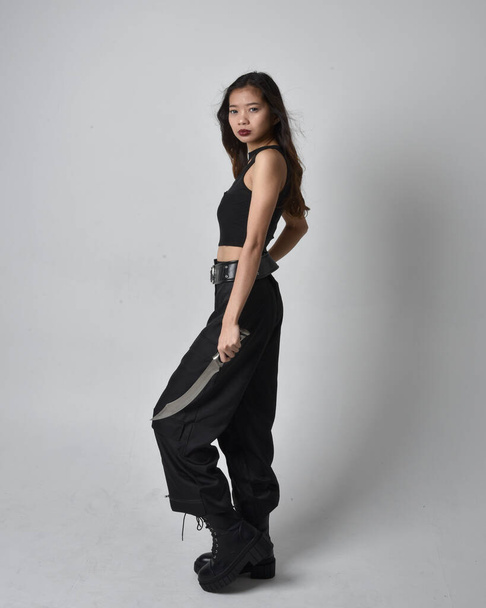 Full length portrait of pretty young asian girl wearing black tank top, utilitarian  pants and leather boots. Standing pose holding a knife,  isolated against a  studio background. - Foto, Imagem