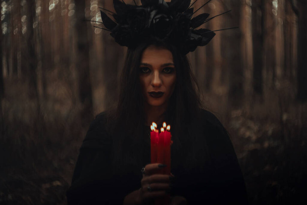 heks in een zwart kostuum houdt kaarsen in een donker bos - Foto, afbeelding