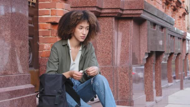 Sfrustrowana młoda Afroamerykanka z plecakiem siedząca na schodach i trzymająca dolary. etniczne czarny tysiąclecia kobieta student stracił karty bankowe, licząc gotówkę, brakuje, wzdycha smutny - Materiał filmowy, wideo