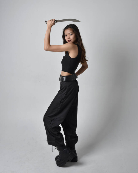 Full length portrait of pretty young asian girl wearing black tank top, utilitarian  pants and leather boots. Standing pose holding a knife,  isolated against a  studio background. - Foto, afbeelding