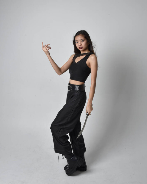 Full length portrait of pretty young asian girl wearing black tank top, utilitarian  pants and leather boots. Standing pose holding a knife,  isolated against a  studio background. - Foto, Bild