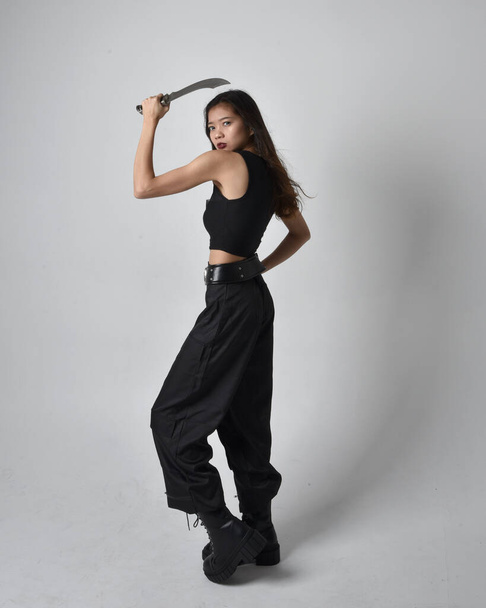 Full length portrait of pretty young asian girl wearing black tank top, utilitarian  pants and leather boots. Standing pose holding a knife,  isolated against a  studio background. - Fotó, kép