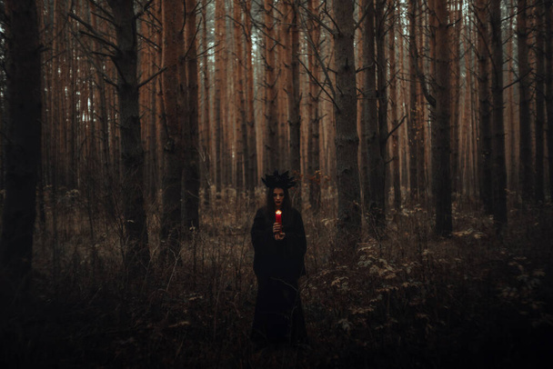 μάγισσα με μαύρη στολή κρατά κεριά σε σκοτεινό δάσος - Φωτογραφία, εικόνα