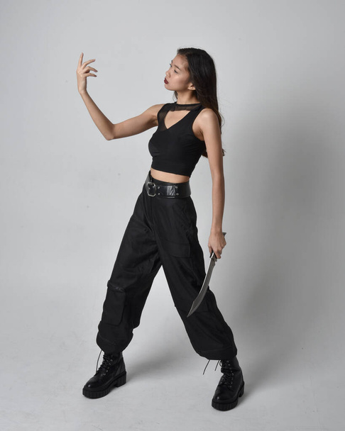 Full length portrait of pretty young asian girl wearing black tank top, utilitarian  pants and leather boots. Standing pose holding a knife,  isolated against a  studio background. - Foto, imagen