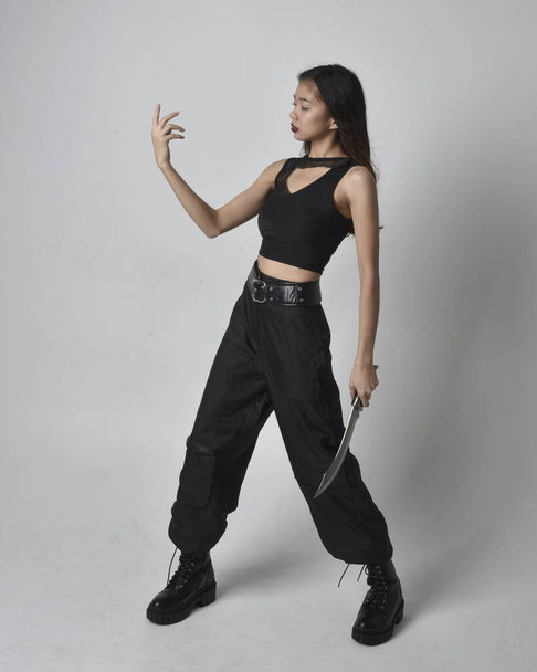 Full length portrait of pretty young asian girl wearing black tank top, utilitarian  pants and leather boots. Standing pose holding a knife,  isolated against a  studio background. - Φωτογραφία, εικόνα