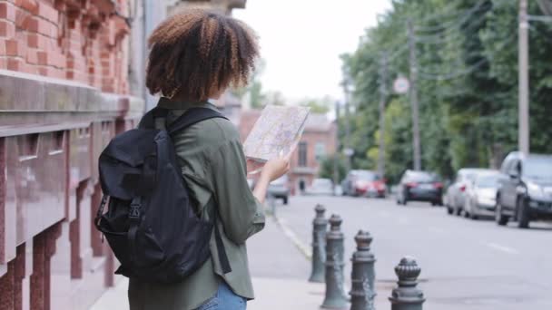 Молода афроамериканська дівчина - туристка з рюкзаком подорожує тільки великим містом, тримаючи паперові карти в руках, перевіряючи напрямок і маршрут планування. Здатність до навігації, незалежна концепція одиночних поїздок - Кадри, відео