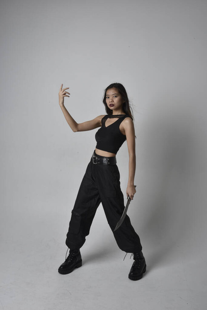 Full length portrait of pretty young asian girl wearing black tank top, utilitarian  pants and leather boots. Standing pose holding a knife,  isolated against a  studio background. - Fotoğraf, Görsel
