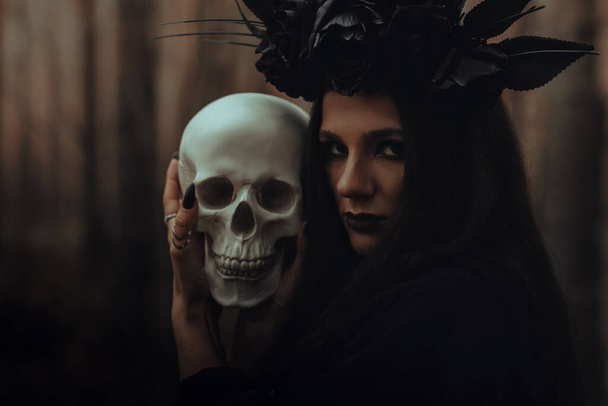 frightening evil witch in black rags holds a dead mans skull in her hands for a dark ritual - Foto, Imagem