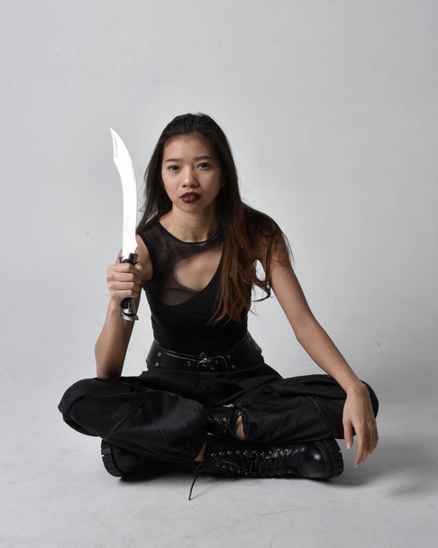 Full length portrait of pretty young asian girl wearing black tank top, utilitarian  pants and leather boots. Sitting pose holding a knife,  isolated against a  studio background. - Zdjęcie, obraz