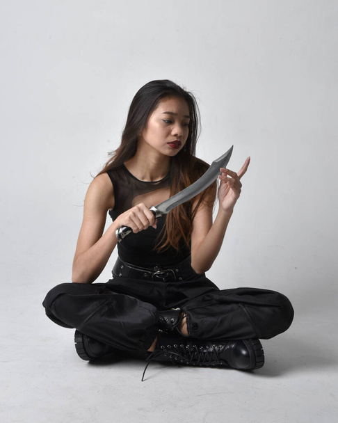 Full length portrait of pretty young asian girl wearing black tank top, utilitarian  pants and leather boots. Sitting pose holding a knife,  isolated against a  studio background. - Fotografie, Obrázek