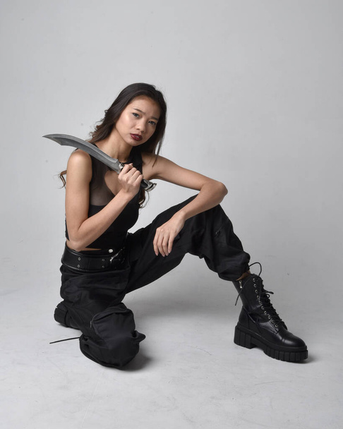Portret młodej Azjatki w czarnej koszulce, użytkowych spodniach i skórzanych butach. Postawa siedząca z nożem, odizolowana od tła pracowni. - Zdjęcie, obraz