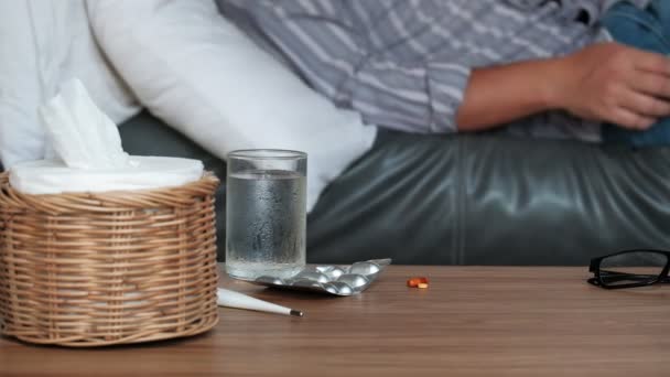 Asiatische männliche Arbeitskräfte fehlen krankheitsbedingt, nehmen die Temperatur mit dem Thermometer, nehmen Medikamente und schlafen zu Hause in Decken auf dem Sofa, Taschentücher und Tabletten auf dem Tisch. - Filmmaterial, Video