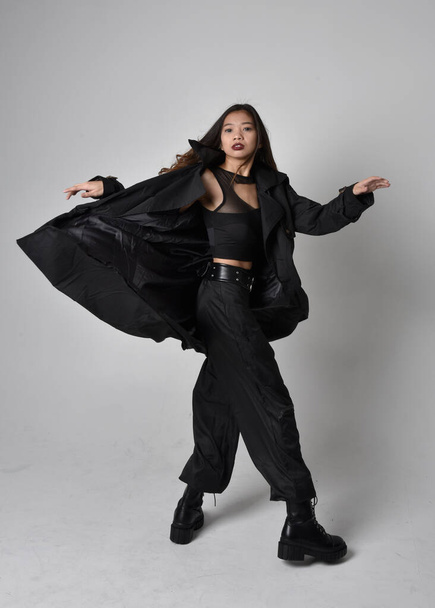 Portret pięknej Azjatki w czarnej koszulce, użytkowych spodniach, skórzanych butach i dużym ciemnym płaszczu. Pozycja stojąca z gestykulacyjnymi ruchami dłoni, odizolowana na tle studia. - Zdjęcie, obraz
