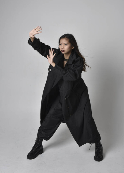 Full length portrait of pretty young asian girl wearing black tank top, utilitarian  pants, leather boots and large dark coat. Standing pose with gestural hand movements,  isolated on  studio background. - Photo, image