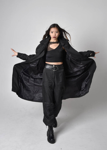 Full length portret van mooie jonge aziatische meisje dragen zwarte tank top, utilitaire broek, lederen laarzen en grote donkere jas. Staande houding met gebaar handbewegingen, geïsoleerd op studio achtergrond. - Foto, afbeelding