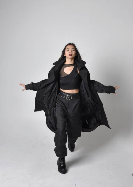 Full length portrait of pretty young asian girl wearing black tank top, utilitarian  pants, leather boots and large dark coat. Standing pose with gestural hand movements,  isolated on  studio background. - Valokuva, kuva
