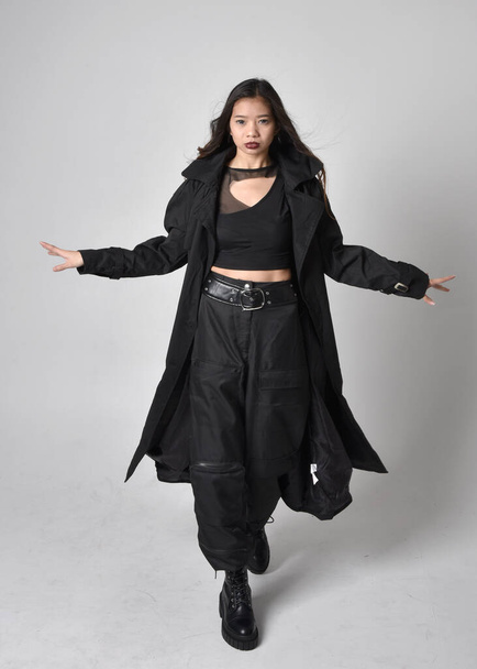 Portret pięknej Azjatki w czarnej koszulce, użytkowych spodniach, skórzanych butach i dużym ciemnym płaszczu. Pozycja stojąca z gestykulacyjnymi ruchami dłoni, odizolowana na tle studia. - Zdjęcie, obraz