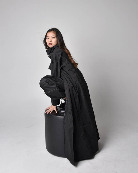 Ganzkörperporträt eines hübschen jungen asiatischen Mädchens mit schwarzem Tank-Top, praktischer Hose, Lederstiefeln und großem dunklen Mantel. Sitzende Pose mit gestischen Handbewegungen, isoliert auf Studiohintergrund. - Foto, Bild