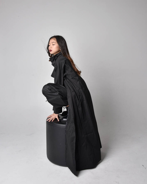 Full length portrait of pretty young asian girl wearing black tank top, utilitarian  pants, leather boots and large dark coat. Sitting pose with gestural hand movements,  isolated on  studio background. - Foto, Bild