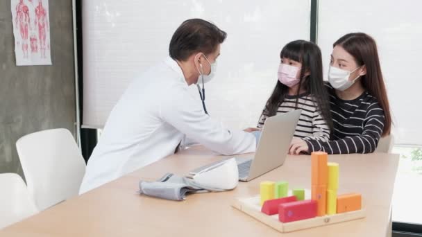 A mother with a face mask brings her sick daughter to an exam appointment with an Asian male doctor at the pediatrics department of a kid's clinic. To treat illnesses and to consult on family health. - Footage, Video