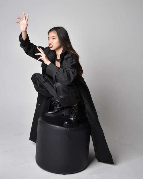 Ganzkörperporträt eines hübschen jungen asiatischen Mädchens mit schwarzem Tank-Top, praktischer Hose, Lederstiefeln und großem dunklen Mantel. Sitzende Pose mit gestischen Handbewegungen, isoliert auf Studiohintergrund. - Foto, Bild