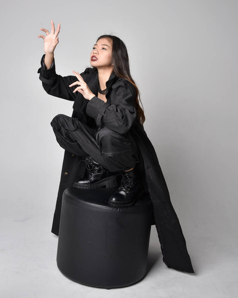 Full length portrait of pretty young asian girl wearing black tank top, utilitarian  pants, leather boots and large dark coat. Sitting pose with gestural hand movements,  isolated on  studio background. - Фото, изображение