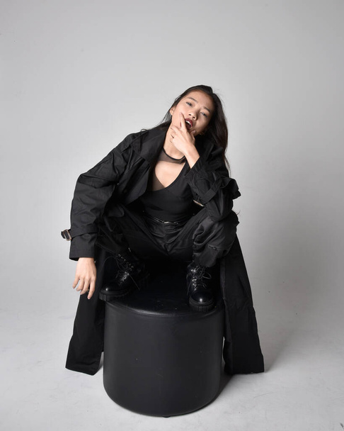 Full length portrait of pretty young asian girl wearing black tank top, utilitarian  pants, leather boots and large dark coat. Sitting pose with gestural hand movements,  isolated on  studio background. - Foto, immagini