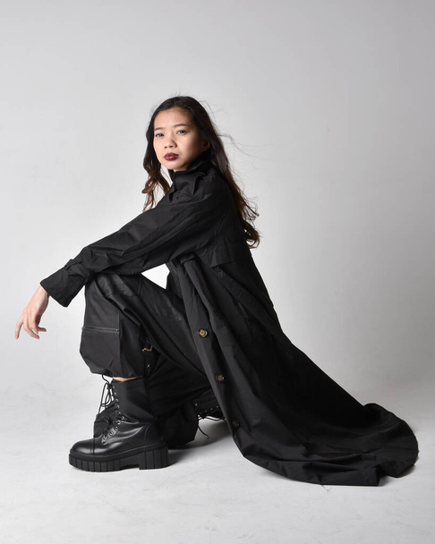 Full length portrait of pretty young asian girl wearing black tank top, utilitarian  pants, leather boots and large dark coat. Sitting pose with gestural hand movements,  isolated on  studio background. - Fotoğraf, Görsel