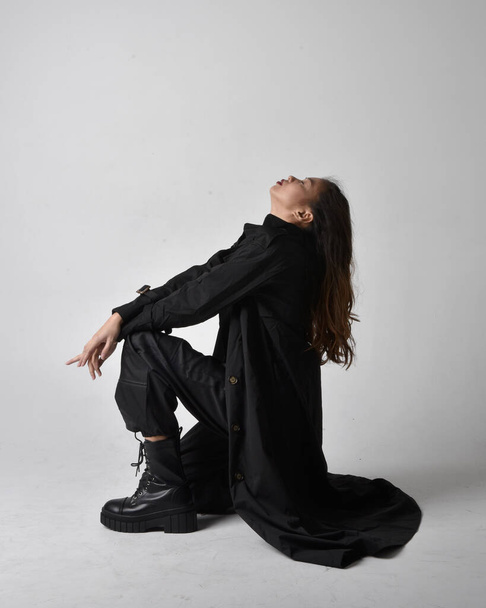 Full length portrait of pretty young asian girl wearing black tank top, utilitarian  pants, leather boots and large dark coat. Sitting pose with gestural hand movements,  isolated on  studio background. - Φωτογραφία, εικόνα
