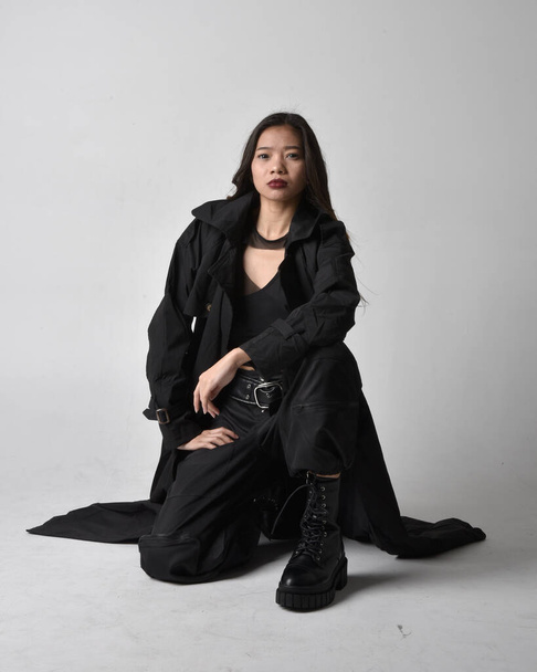 Full length portrait of pretty young asian girl wearing black tank top, utilitarian  pants, leather boots and large dark coat. Sitting pose with gestural hand movements,  isolated on  studio background. - Fotografie, Obrázek