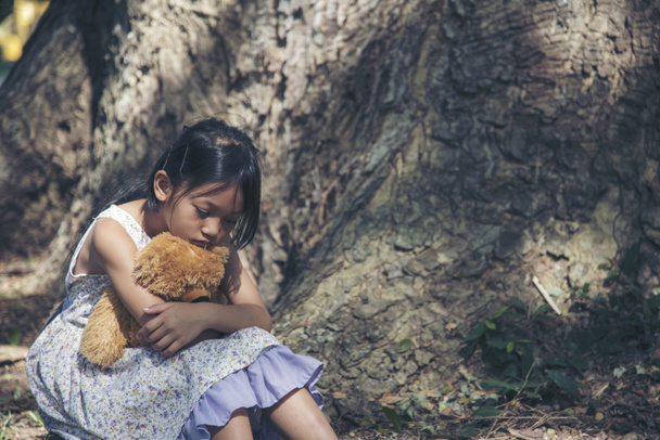 Trauriges Mädchen umarmt Teddybär sitzt unter Baum Traurigkeit allein im grünen Park. Einsames Mädchen traurig unglücklich sitzen draußen umarmen beste Freundin Spielzeug. Autismus Kind spielen Teddybär besten Freund. Gewalt in der Familie - Foto, Bild