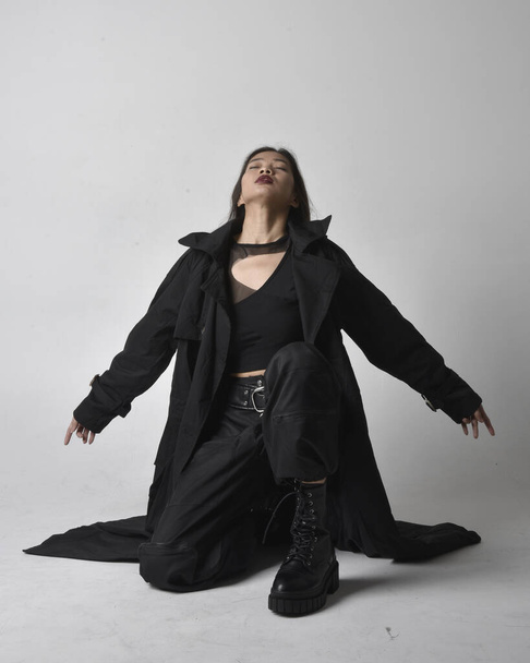 Full length portrait of pretty young asian girl wearing black tank top, utilitarian  pants, leather boots and large dark coat. Sitting pose with gestural hand movements,  isolated on  studio background. - Photo, Image