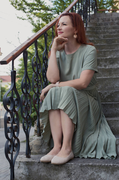 Young red longhair woman dressing green dress sitting brooding on the stairs - Фото, изображение
