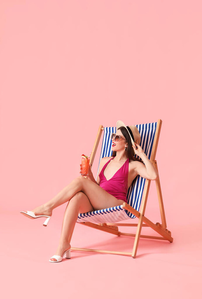 Beautiful young woman with cocktail sitting on deck chair against color background - Photo, Image