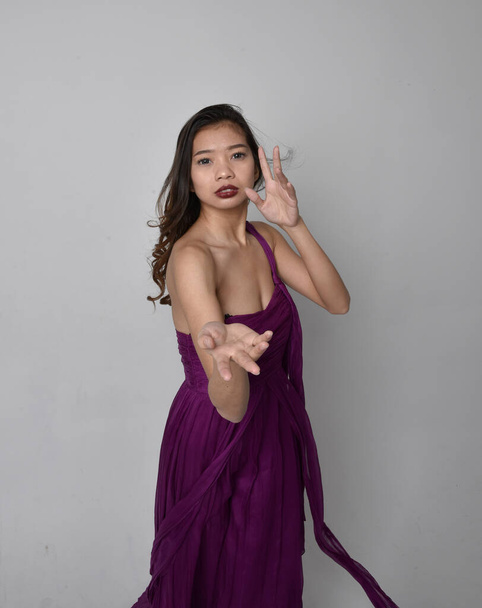 Close up portrait of pretty brunette asian girl wearing purple flowing  gown. Gestural hand movements on grey studio background. - Foto, imagen