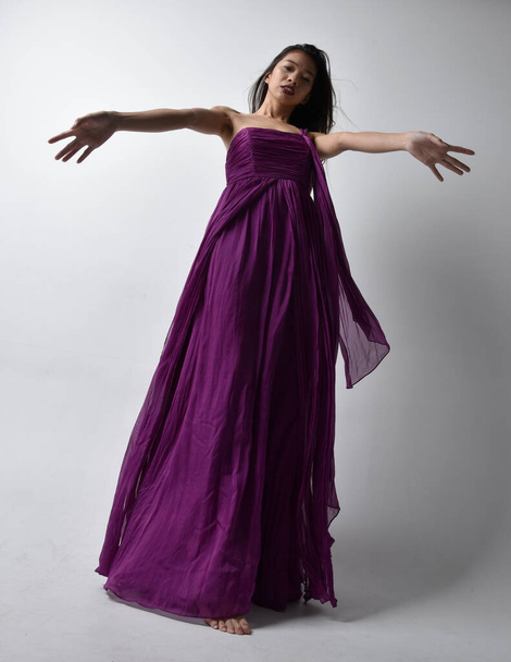 Full length  portrait of pretty brunette asian girl wearing purple flowing  gown. Standing pose in low angle on on studio background. - Foto, Imagem