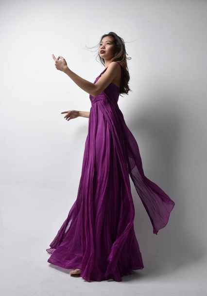 Full length  portrait of pretty brunette asian girl wearing purple flowing  gown. Standing pose in low angle on on studio background. - Foto, Imagen