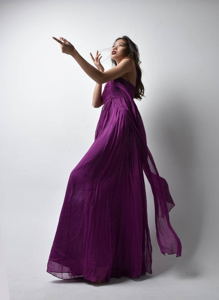 Full length  portrait of pretty brunette asian girl wearing purple flowing  gown. Standing pose in low angle on on studio background. - Fotó, kép