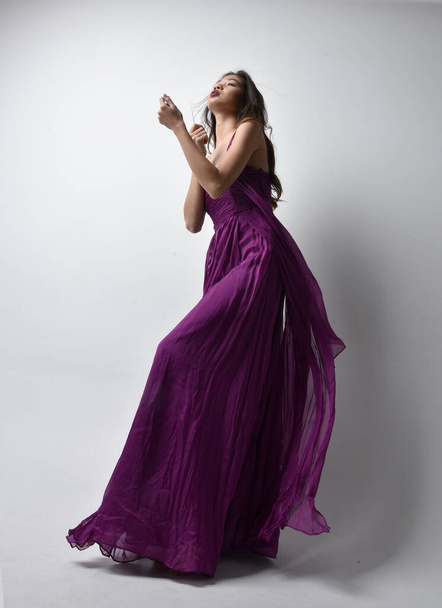 Full length  portrait of pretty brunette asian girl wearing purple flowing  gown. Standing pose in low angle on on studio background. - Φωτογραφία, εικόνα