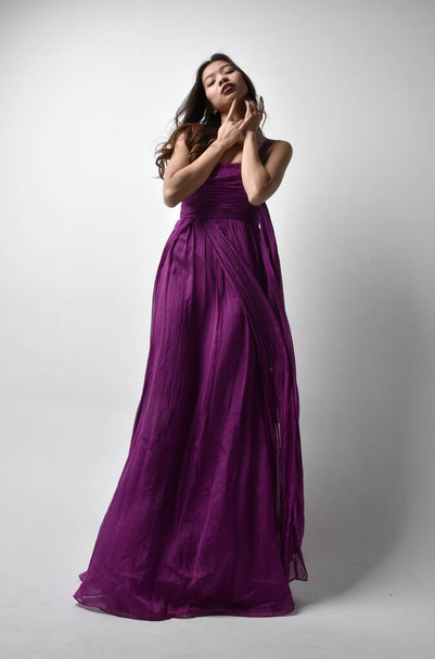 紫の流れるガウンを着てかなりブルネットのアジアの女の子の完全な長さの肖像画。低い角度で立ちポーズでスタジオ背景. - 写真・画像