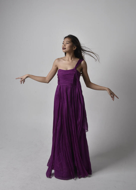 Full length  portrait of pretty brunette asian girl wearing purple flowing  gown. Standing, dancing pose on on studio background. - Foto, Imagen
