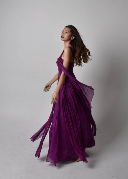 Full length  portrait of pretty brunette asian girl wearing purple flowing  gown. Standing, dancing pose on on studio background. - Фото, зображення