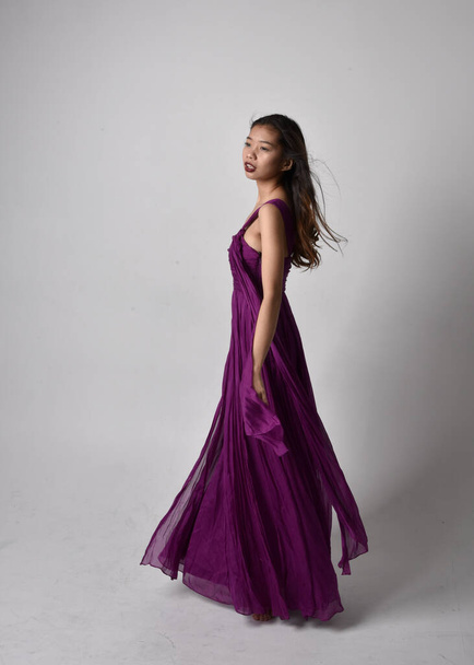 Full length  portrait of pretty brunette asian girl wearing purple flowing  gown. Standing, dancing pose on on studio background. - Foto, immagini