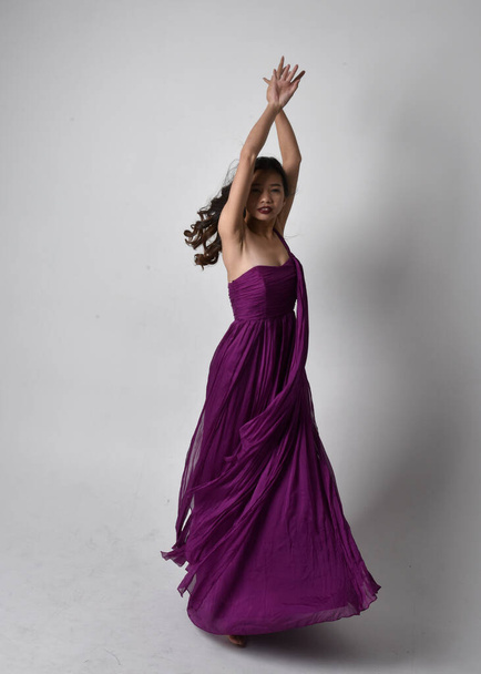 Full length  portrait of pretty brunette asian girl wearing purple flowing  gown. Standing, dancing pose on on studio background. - Fotoğraf, Görsel