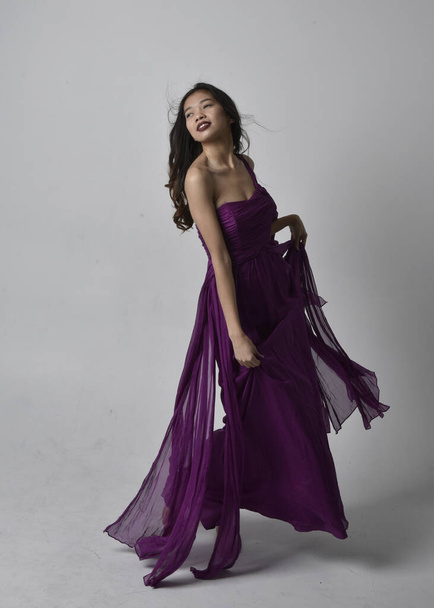 Full length  portrait of pretty brunette asian girl wearing purple flowing  gown. Standing, dancing pose on on studio background. - Photo, Image