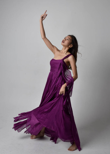 Full length  portrait of pretty brunette asian girl wearing purple flowing  gown. Standing, dancing pose on on studio background. - Photo, image
