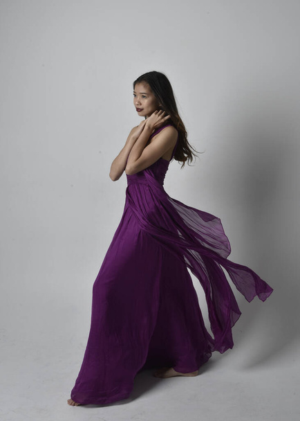 Full length  portrait of pretty brunette asian girl wearing purple flowing  gown. Standing, dancing pose on on studio background. - Zdjęcie, obraz