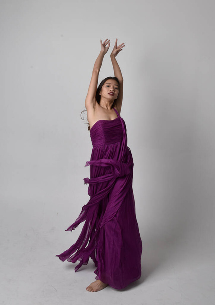 Full length  portrait of pretty brunette asian girl wearing purple flowing  gown. Standing, dancing pose on on studio background. - 写真・画像