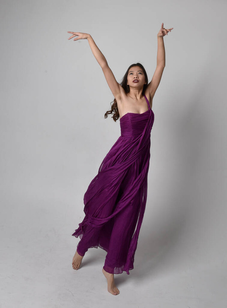 Full length  portrait of pretty brunette asian girl wearing purple flowing  gown. Standing, dancing pose on on studio background. - Foto, Imagen